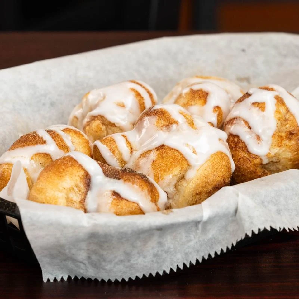 Golden baked rolls drizzled with white icing sit in a parchment-lined basket on a dark wooden table.
